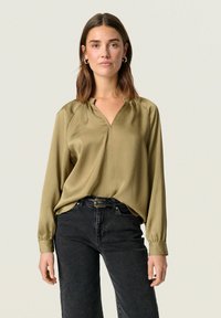 Olive green, long-sleeved blouse with a v-neck and textured surface. Paired with high-waisted black jeans and a silver belt buckle.