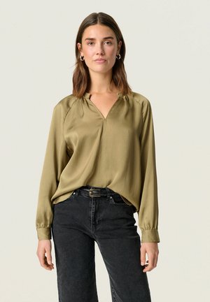Olive green, long-sleeved blouse with a v-neck and textured surface. Paired with high-waisted black jeans and a silver belt buckle.