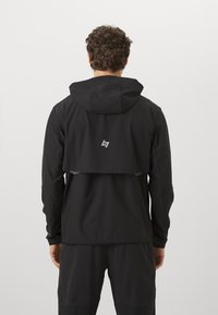 Μαύρο ελαφρύ μπουφάν με κουκούλα, με διακοσμητική ανακλαστική λεπτομέρεια στο πίσω μέρος και ελαστικές μανσέτες. Λείο, αδιάβροχο υλικό.