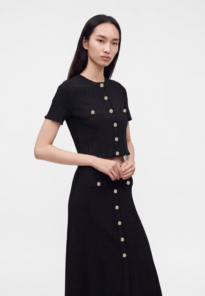 Femme portant un crop top noir à manches courtes boutonné et une jupe taille haute assortie avec des boutons dorés, debout devant un fond clair uni.