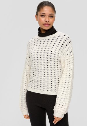 Weißer Strickpullover mit offener Webstruktur, weiten Ärmeln und gerippten Rändern, getragen über einem schwarzen Rollkragenpullover, kombiniert mit schwarzen gerippten Hosen.