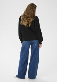 Schwarzer Pullover mit elastischen Bündchen und weiter Passform, kombiniert mit hoch taillierter, weit geschnittener blauer Jeans aus Denim. Weiße Leinenschuhe sind sichtbar.