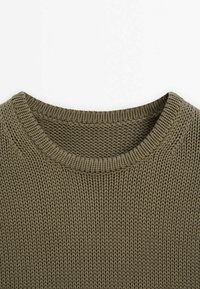 Gros plan sur l'encolure d'un pull tricoté vert kaki avec un col côtelé rond et un motif de tricot texturé.