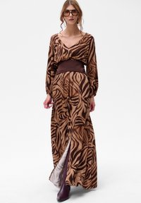 Sukienka maxi w kolorze brązowym i beżowym w wzór zebry, z długimi rękawami, głębokim dekoltem w kształcie litery V oraz szerokim brązowym paskiem w talii. Tekstura wydaje się gładka.