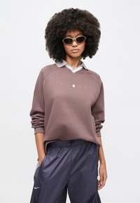 Καφέ φούτερ με γκρι γιακά, με v-λαιμόκοψη και λογότυπο. Συνδυάζεται με σκούρο, φαρδύ παντελόνι με ορατό λογότυπο μάρκας.