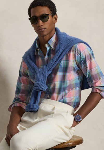 Light blue knit sweater draped over a colorful plaid shirt, cream pleated trousers, and a blue watch with a brown leather strap. Dark sunglasses.
