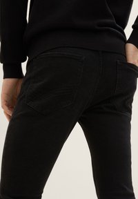 Jeans de mezclilla negra con un corte ajustado, que presentan dos bolsillos traseros y un detalle de costura sutil. La tela tiene una textura suave y un acabado mate.