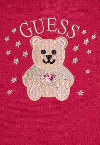Un tessuto rosso presenta un orso ricamato con un cuore rosa di paillettes, circondato da stelle argentate e dalla parola "GUESS" sopra.