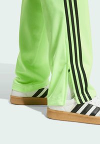 Neon green track pants with black side stripes and zippered hems, paired with white sneakers featuring black stripes and gum soles.