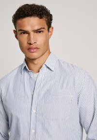 Camisa de botones a rayas azul y blanca con un bolsillo, hecha de tela de algodón. Presenta un cuello con solapa y detalles de botones en contraste.