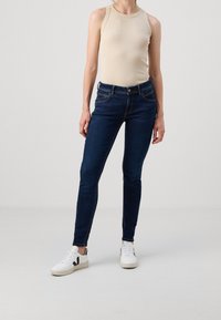 Mörkblå denimjeans med slim fit, har midjehöjd, fem fickor och kontrasterande sömmar. Matchas med en beige top och vita sneakers.