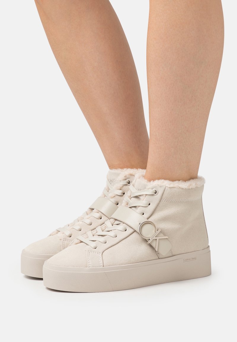 Beżowe sneakersy typu high-top z zamszu z miękkim futrzanym mankietem. Posiadają sznurowaną przód, płaską podeszwę oraz okrągły akcent logo na pasku.