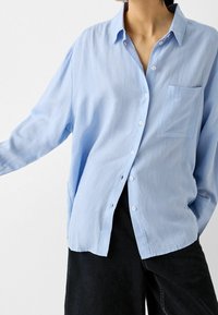Ljusblå skjorta med knäppning och långa ärmar, en bröstficka och avslappnad passform. Tillverkad av ett mjukt, lättviktigt material.