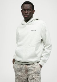 Helle graue Kapuzen sweatshirt aus weichem Material mit einer Kängurutasche und subtilen Logo-Details. Getragen mit Camouflage-muster Hosen.