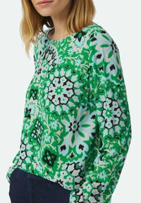 Groene blouse met een patroon van bloemen, met zwarte en lichtblauwe accenten. Lange mouwen en een ronde halslijn. Glad stoftextuur.