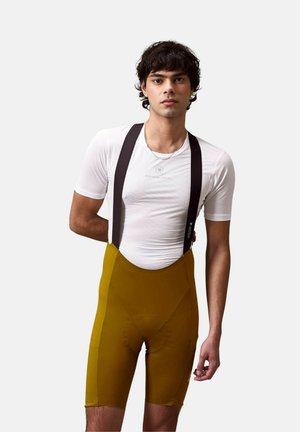Jonge man die een wit shirt met korte mouwen en mosterdgele wielerbroek met zwarte bretels draagt, staand tegen een effen achtergrond.