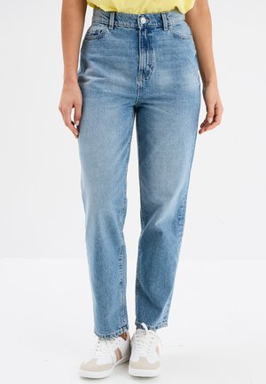 Persona che indossa jeans azzurri a vita alta, top giallo e sneakers bianche in piedi con le braccia rilassate lungo i fianchi.