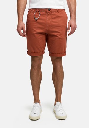 Mężczyzna w rdzawej, podwiniętej szortach z plecionym brelokiem i białych trampkach na białym tle.