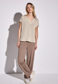 Světlé béžové tričko s krátkým rukávem a výstřihem do V, kombinované s volnými hnědými kalhotami a světle barevnými teniskami. Hladká textilie.