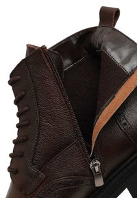 Botte en cuir marron foncé avec une fermeture éclair latérale partiellement ouverte et des lacets bruns sur le côté extérieur.