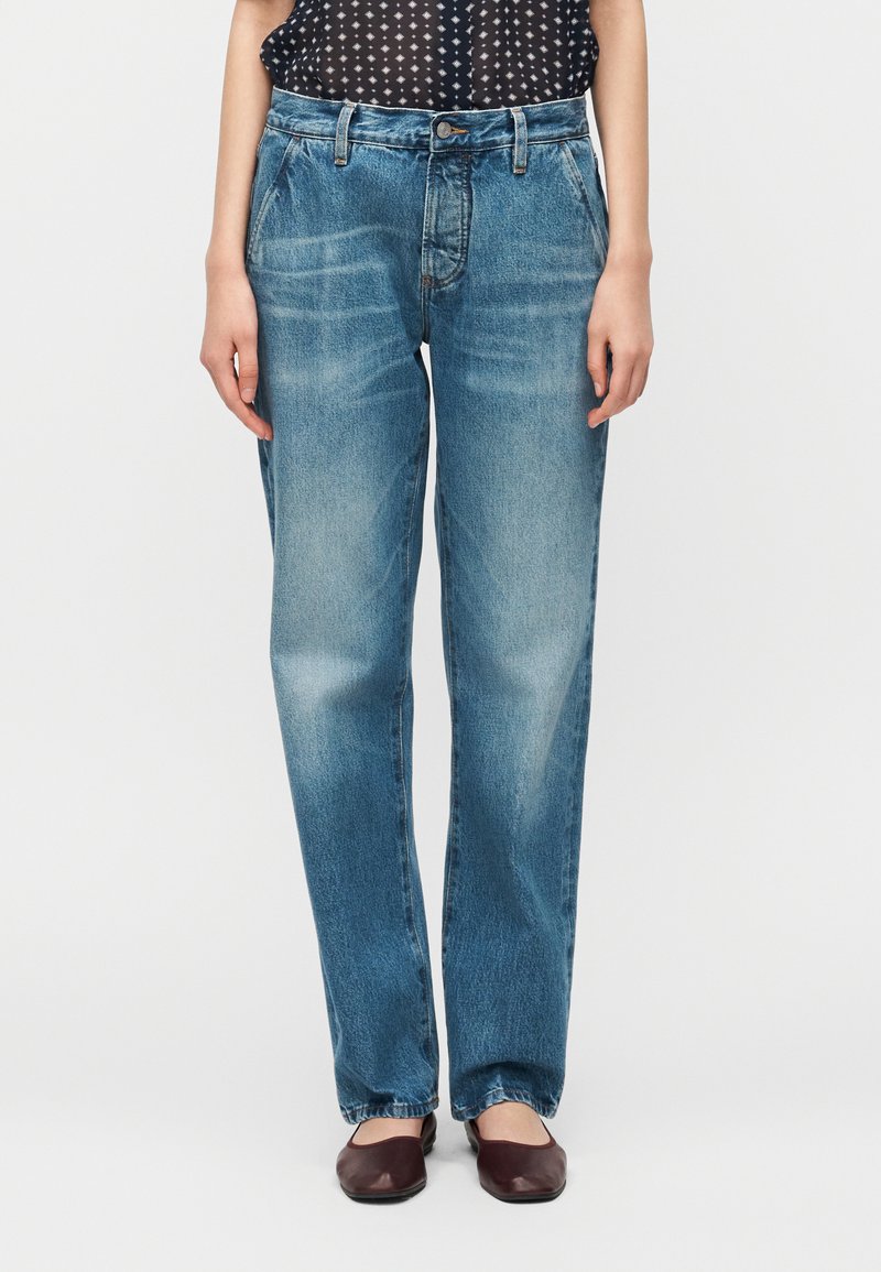 Άτομο που φοράει φαρδιές, ξεθωριασμένες μπλε τζιν παντελόνες με μπροστινές τσέπες, σε συνδυασμό με μαύρο αμάνικο τοπ με σχέδια και σκούρα καφέ επίπεδα παπούτσια.