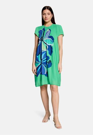 Femme debout portant une robe vert vif longueur genou avec un grand imprimé floral bleu, manches courtes et talons ouverts beige sur fond uni.
