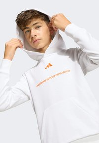 Biela mikina vyrobená z mäkkého materiálu, s oranžovým logom "ADIDAS SPORTSWEAR" a tromi oranžovými pruhmi na prednej strane. Obsahuje predné vrecko.