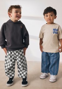 Dve otroci stojita drug ob drugem. Eden nosi temno sivo kapuco, črno-bele karo hlače in črne superge. Drugi ima bež majico z modrim napisom in svetlo modre črtaste hlače ter bele superge.