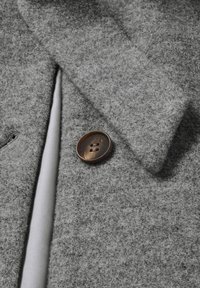 Grauer Filzstoff mit glatter Textur, versehen mit einem braunen Knopf mit zwei Löchern. Eine Seite ist mit einem Satinabschluss veredelt.