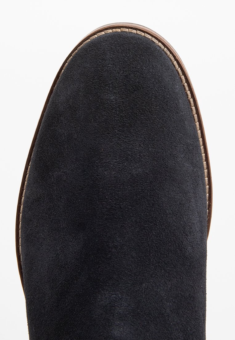 Schwarze Wildlederstiefel mit abgerundetem Zeh und sichtbaren Nähten entlang der Sohle. Glatte Textur, minimalistisches Design und eine flache, braune Sohle.