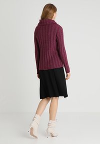 Maglione a coste bordeaux con collo alto a cowl, abbinato a una gonna nera e stivali alla caviglia morbidi color crema. Design semplice e aderente.