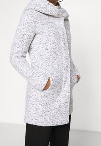 Cappotto grigio chiaro, strutturato, con collo a imbuto, caratterizzato da due tasche laterali e un taglio dritto. Il tessuto presenta fibre nere e bianche.