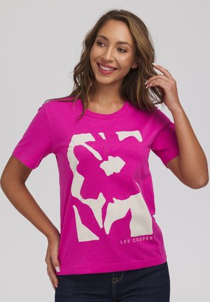 Uśmiechnięta kobieta z długimi włosami, ubrana w jaskraworóżową koszulkę Lee Cooper z białym abstrakcyjnym kwiatowym wzorem, pozująca z jedną ręką przy włosach.