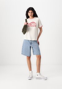 Cotton T-shirt in cream with red trim and logo, paired with faded denim shorts. White high-top sneakers complete the look.