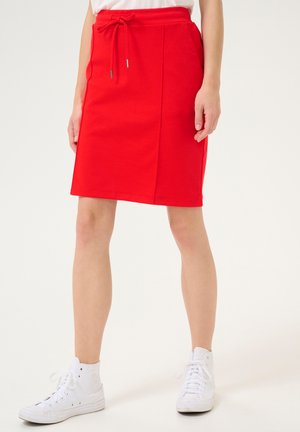 Fustă roșie din bumbac cu talie cu șnur, până la genunchi, cu cusături verticale. Purtează adidași albi cu glezna înaltă. Design simplu, casual.