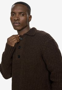 Barna kötött pulóver, texturált mintával, elöl gombos záródással és gallérral. Az anyag vastagnak és melegnek tűnik.