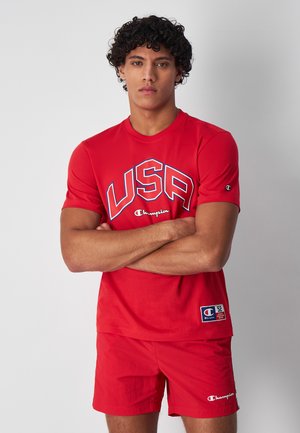 Camiseta de algodón roja con "USA" en texto azul y blanco, con un logo de Champion, combinada con pantalones cortos rojos. Diseño simple y ajuste atlético.