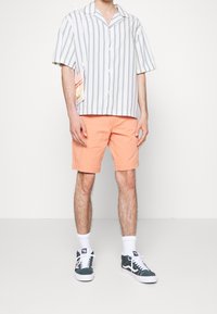 Chemise à manches courtes boutonnée avec des rayures verticales bleues et blanches, associée à un short pêche et des baskets vertes. Comprend un petit accessoire sac.