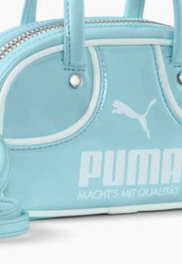 Svetlo modra lakirana torbica z belimi detajli, okrogle oblike, zadrgo za zapiranje, kratkimi ročaji in izstopajočim belim logotipom "PUMA".