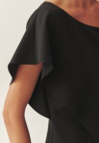 Fekete ruha lebegő, aszimmetrikus nyakkivágással és fodros ujjal. Simább textúra és testhezálló kialakítás, amely kiemeli az elegáns sziluettet.