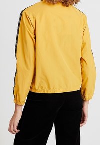 Żółta lekka kurtka z okrągłym dekoltem, elastycznym dołem i czarnymi pasami po bokach z białym tekstem. Teksturowana, gładka tkanina.