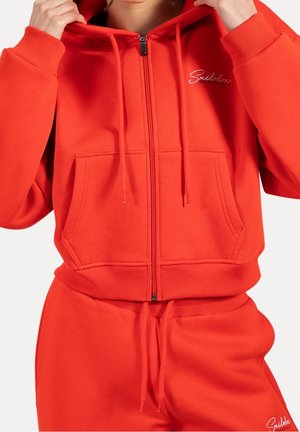 Person trägt einen leuchtend roten Kapuzenpullover mit Reißverschluss und passende Jogginghose mit Kordeln, Hände justieren die Kapuze, kleines weißes Logo auf Brust und Hose.