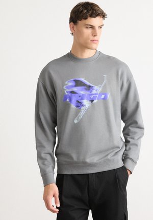 Young man wearing a grey sweatshirt with a blue abstract graphic and the word "PRIGO," paired with black pants, standing against white background.