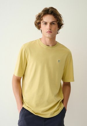 Junger Mann mit lockigem Haar, der ein hellgelbes T-Shirt mit kleinem Palmenlogo auf der Brust trägt, Hände in den Taschen einer dunklen Hose, neutraler Hintergrund.
