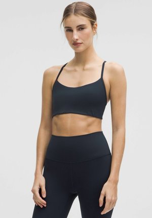 Kvinde med håret bundet tilbage iført en sort sports-bh og sorte høj-taljede leggings, stående mod en ensfarvet lys baggrund.