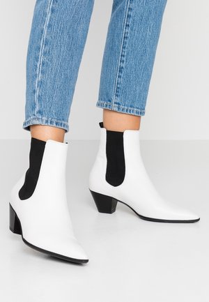 Bottines blanches à la cheville avec panneaux latéraux élastiques noirs et talons épais, portées avec un jean bleu clair raccourci sur un fond uni clair.