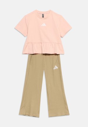 Ροζ κοντομάνικη μπλούζα με βολάν στο τελείωμα και μπεζ παντελόνι φαρδιά με ρίγες, και τα δύο με λευκό λογότυπο Adidas σε ανοιχτό φόντο.