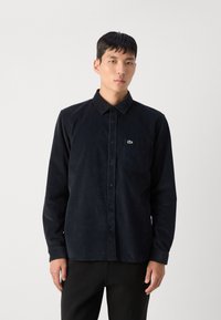 Zwart corduroy knoop-overhemd met een kraag, een borstzak en een klein groen logo. Model draagt het overhemd met zwarte broek.