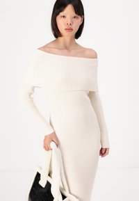Rebrasta haljina s dugim rukavima, off-the-shoulder, u boji krem. Mekan materijal s prilagođenim krojem. Crna torba s bijelim umjetnim krznom.