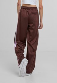 Pantalones deportivos anchos y marrones con rayas laterales blancas y rosas, una cinturilla y un bolsillo trasero. Combinados con zapatillas blancas.